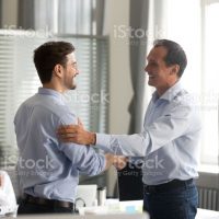 Smiling middle-aged ceo promoting motivating worker shaking hands congratulating with achievement promising respect bonus thanking for good work, team applauding, employee reward recognition concept
